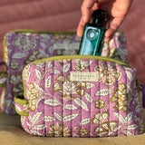 Trousse de toilette Carthage Lilas ouverte avec un flacon, sur un coussin rose
