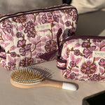 Trousses de toilette Carthage Prune posées sur un coussin, avec une brosse à cheveux