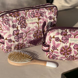 Trousses de toilette Carthage Prune posées sur un coussin, avec une brosse à cheveux