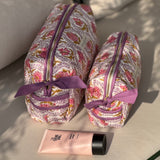 Trousses de toilette Éphèse Rose à nœud lilas posées sur un banc avec une crème