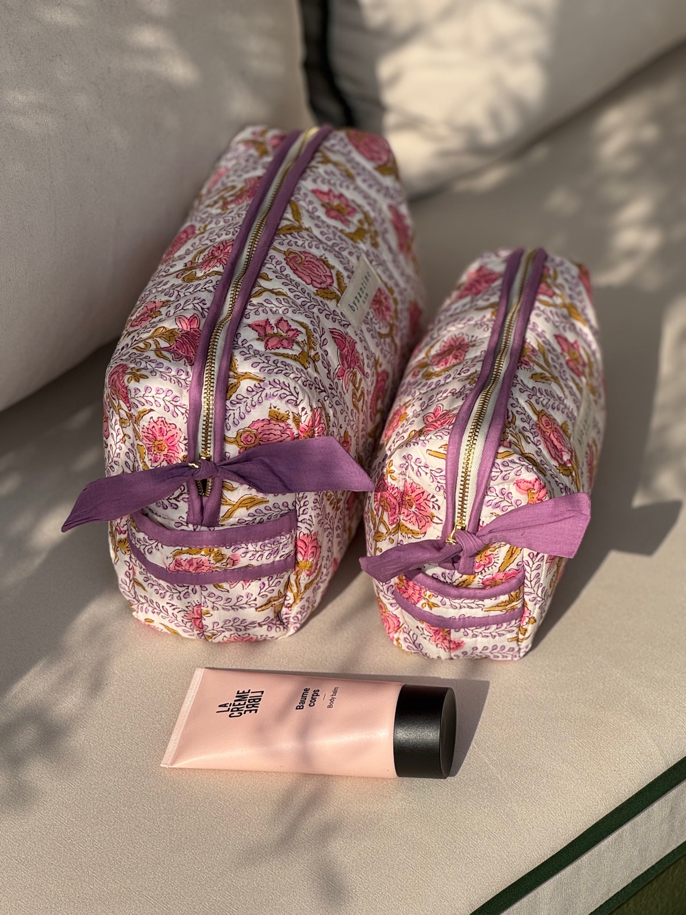 Trousses de toilette Éphèse Rose à nœud lilas posées sur un banc avec une crème