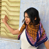 Grand foulard Séléné Fauve porté sur les épaules devant un mur bleu, bordure rose et bleu roi