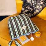 Grande trousse beauté Flora Bleu marine couchée sur un fauteuil moutarde, avec des soins
