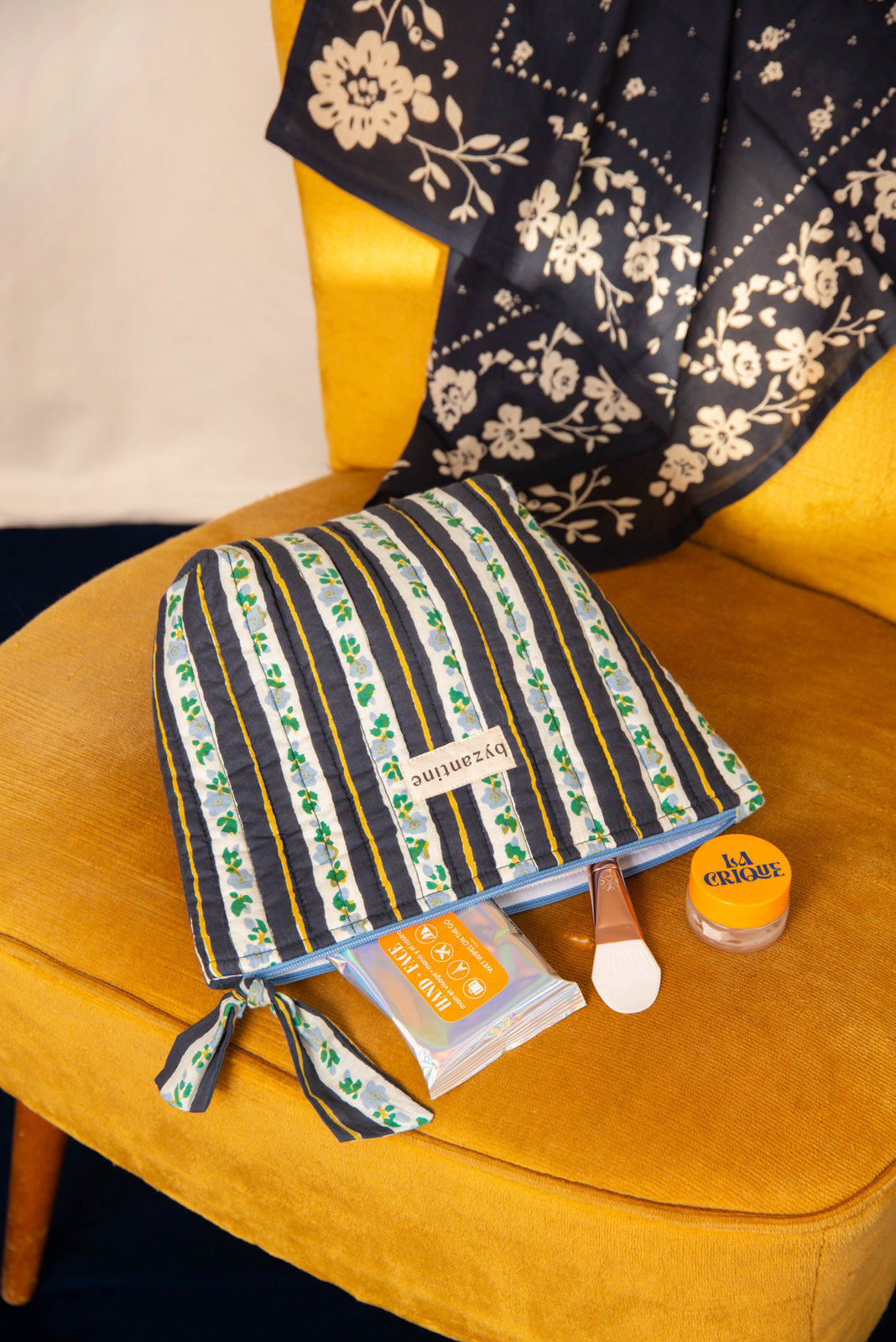 Grande trousse beauté Flora Bleu marine couchée sur un fauteuil moutarde, avec des soins