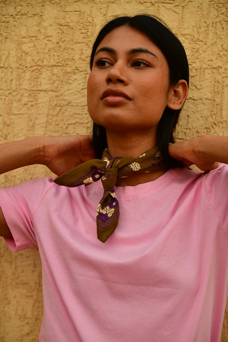Petit foulard Anastase Olive noué au cou, porté avec un tee-shirt rose devant un mur jaune