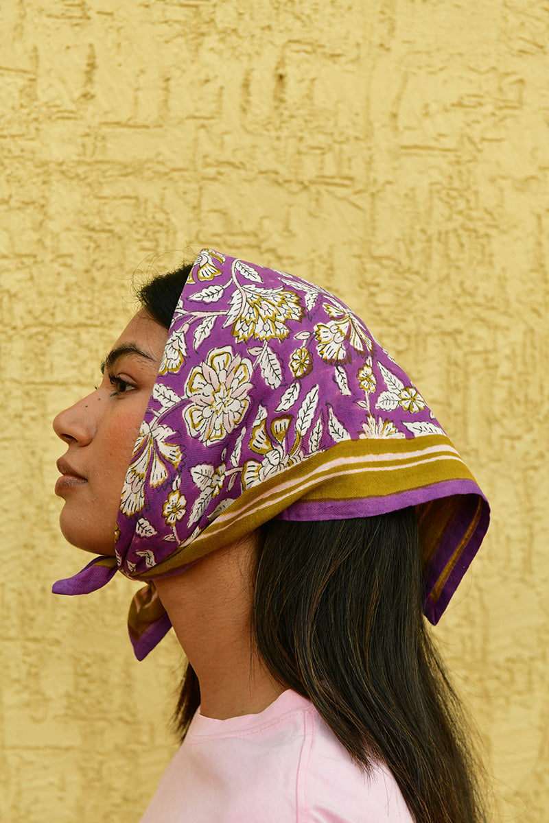 Petit foulard Carthage Violet porté en fichu sur la tête, devant un mur jaune