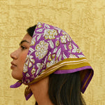 Petit foulard Carthage Violet porté en fichu sur la tête, devant un mur jaune