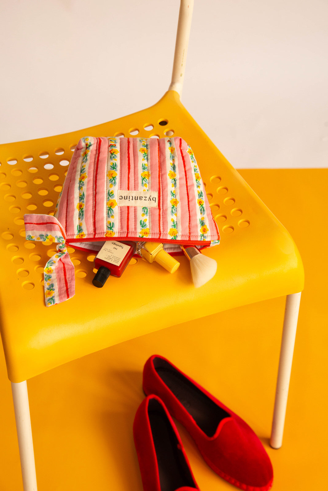 Petite trousse beauté Flora Rose à nœud ouverte sur une chaise jaune, soins éparpillés et ballerines rouges
