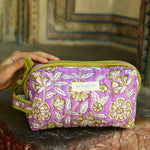 Petite trousse de toilette Carthage Lilas posée sur un muret, main sur la trousse