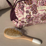 Trousse de toilette Carthage Prune posée à côté d'une brosse à cheveux