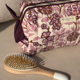 Trousse de toilette Carthage Prune posée sur un canapé, avec une brosse à cheveux
