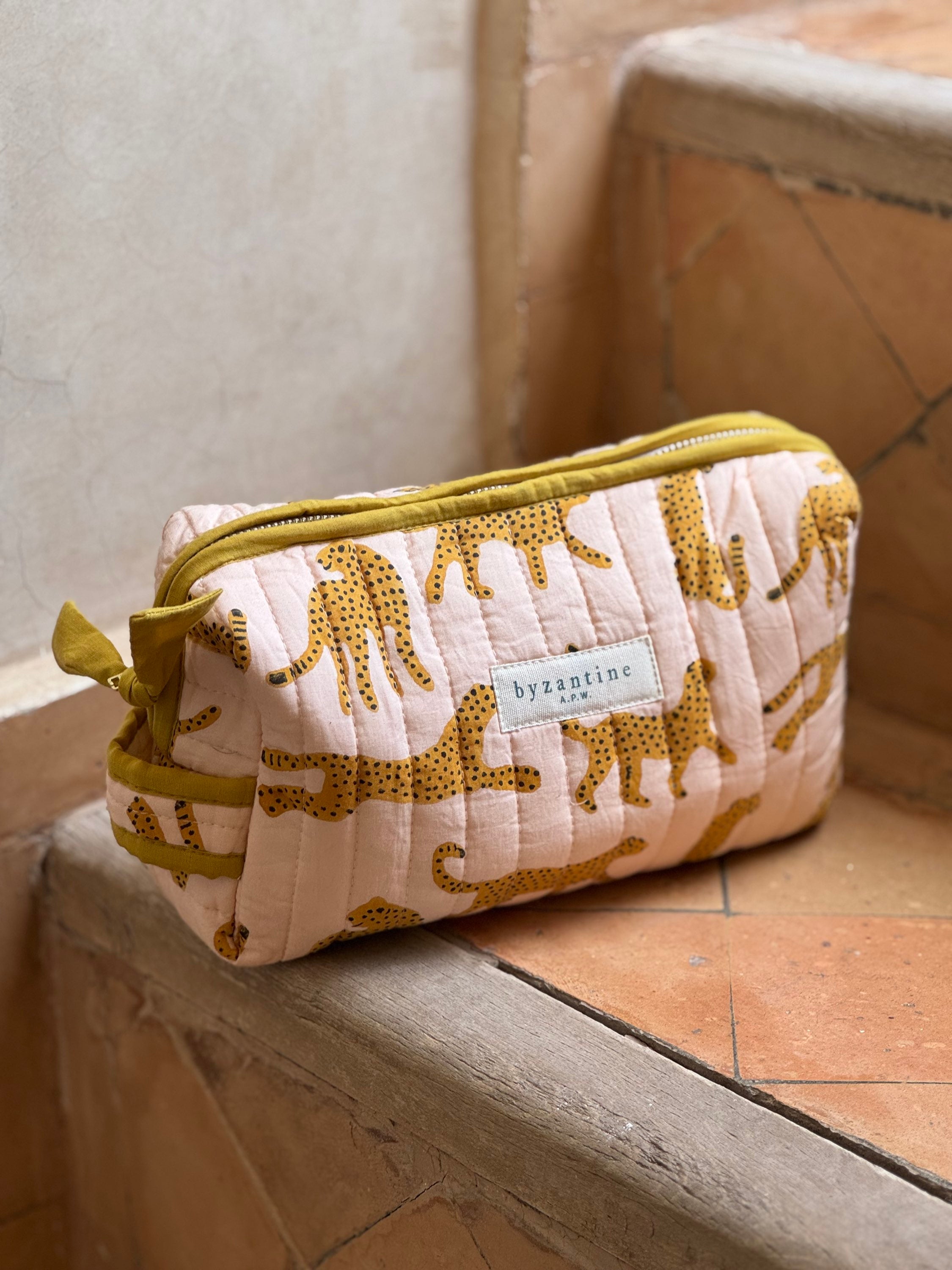 Petite trousse de toilette Fauve Rose à passepoil moutarde posée sur un banc