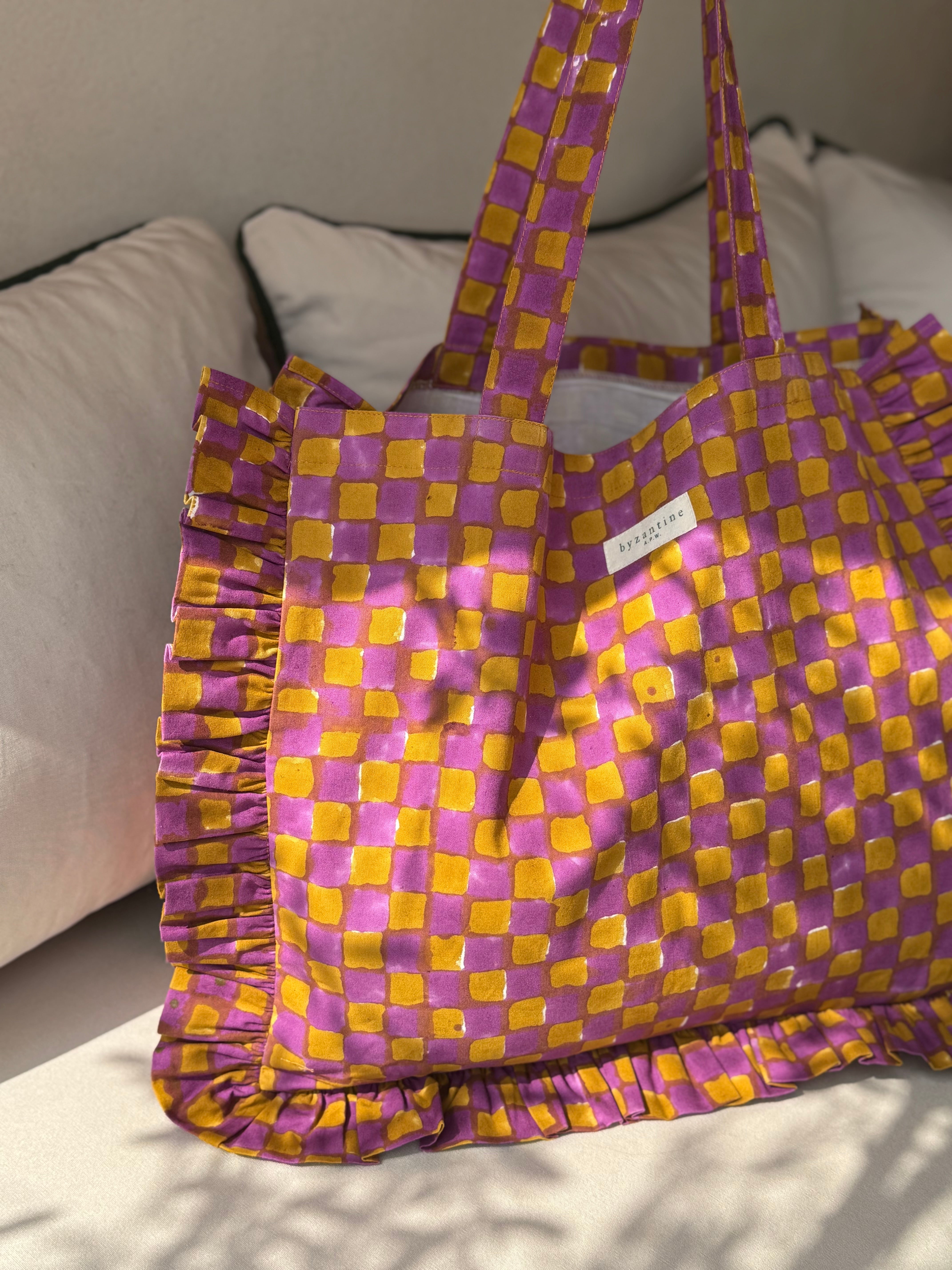 Sac froufrou Moutarde violet à damier, anses longues, étiquette Byzantine, posé au soleil sur un canapé