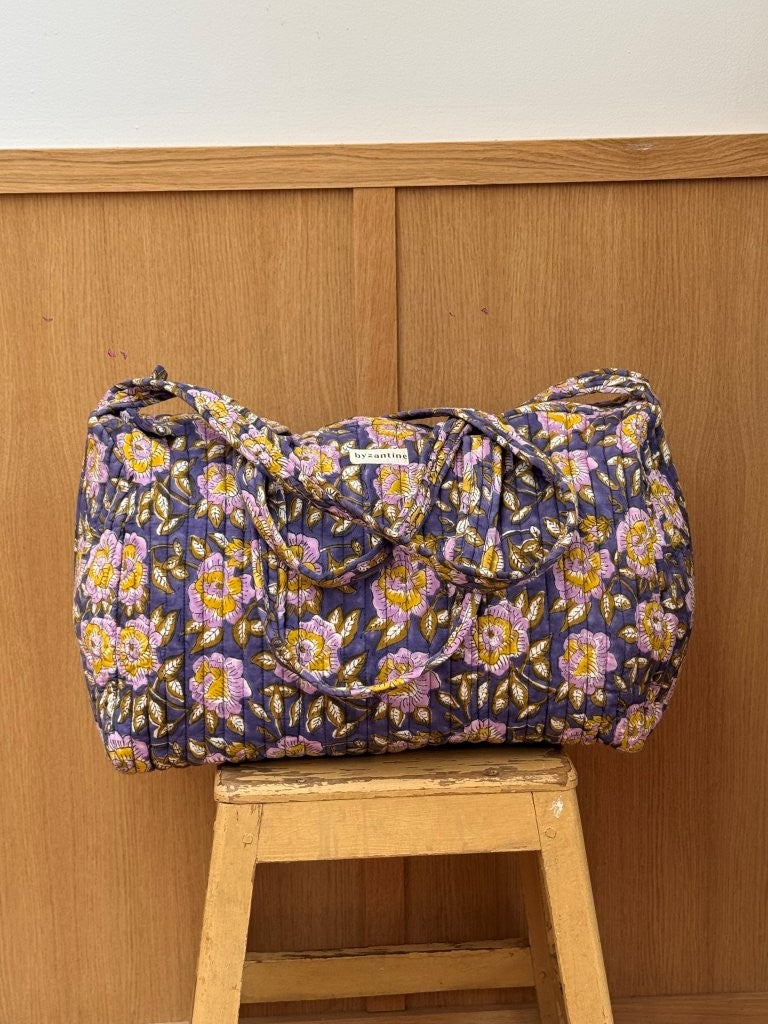 Sac weekend matelassé N°113 Bleu de Byzantine, fleurs jaunes et lilas sur fond bleu, posé sur un tabouret en bois