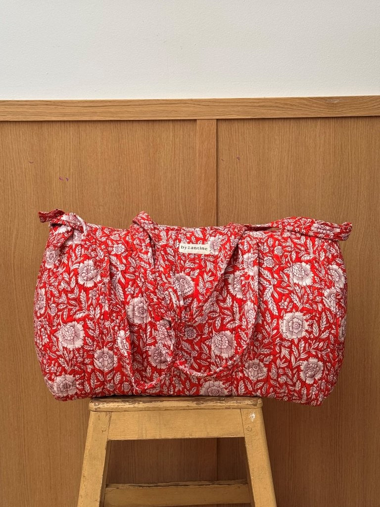 Sac weekend matelassé Antioche Tomato de Byzantine, fleurs blanches sur fond rouge, posé sur un tabouret en bois