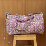 Sac weekend matelassé Carthage Lilas de Byzantine, imprimé fleuri lilas, posé sur un tabouret en bois