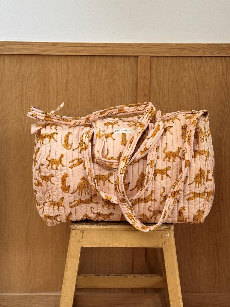 Sac weekend matelassé Fauve Rose de Byzantine, guépards ocre sur fond rose pâle, posé sur un tabouret en bois