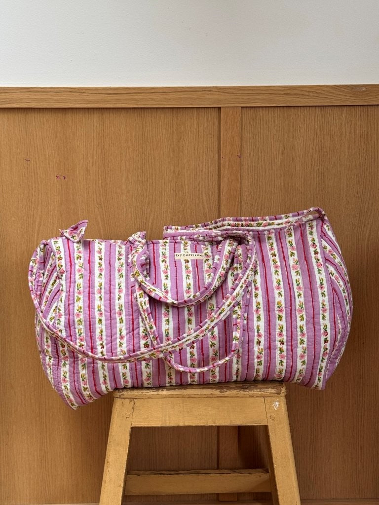 Sac weekend matelassé Flora Lilas de Byzantine, rayures fleuries rose et violet, posé sur un tabouret en bois