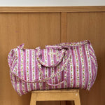 Sac weekend matelassé Flora Lilas de Byzantine, rayures fleuries rose et violet, posé sur un tabouret en bois