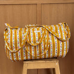 Sac weekend matelassé Flora Passion de Byzantine, rayures fleuries jaune moutarde, posé sur un tabouret en bois