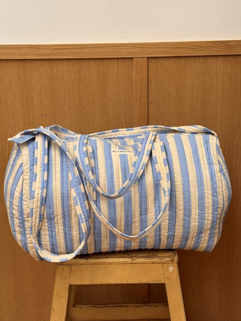 Sac weekend matelassé Pacha Bleu ciel de Byzantine, rayures bleu ciel et écru, posé sur un tabouret en bois
