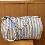 Sac weekend matelassé Pacha Bleu ciel de Byzantine, rayures bleu ciel et écru, posé sur un tabouret en bois