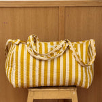 Sac weekend matelassé Pacha Passion de Byzantine, rayures jaune moutarde et blanches, posé sur un tabouret en bois