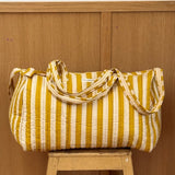 Sac weekend matelassé Pacha Passion de Byzantine, rayures jaune moutarde et blanches, posé sur un tabouret en bois