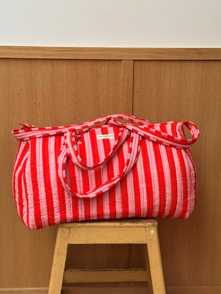 Sac weekend matelassé Pacha Tomato de Byzantine, rayures rouges et roses, posé sur un tabouret en bois