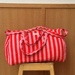 Sac weekend matelassé Pacha Tomato de Byzantine, rayures rouges et roses, posé sur un tabouret en bois