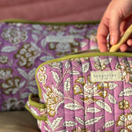 Main glissant une brosse à dents dans la petite trousse Carthage Lilas, devant la trousse de toilette XL