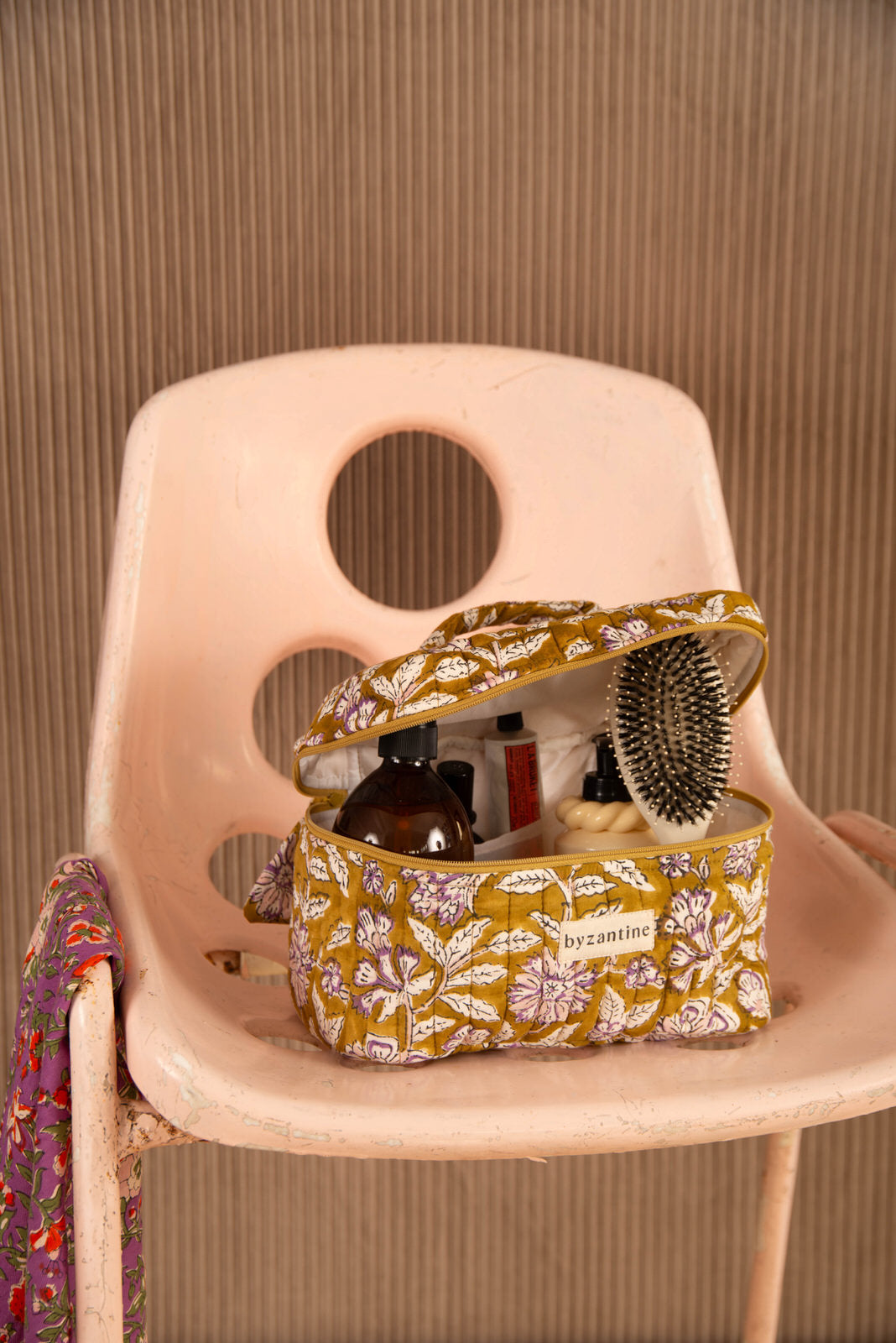 Trousse vanity Carthage Kaki ouverte, avec brosse et flacons, sur une chaise rose