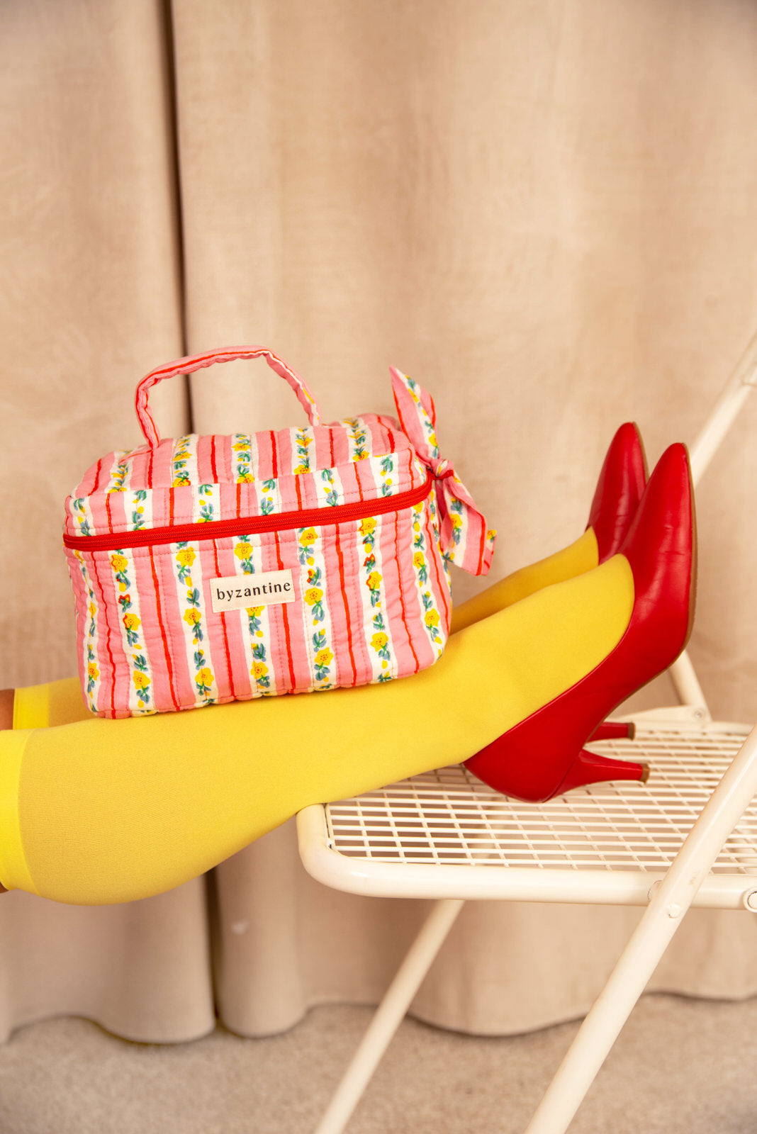 Trousse vanity Flora Rose posée sur des jambes en collants jaunes, escarpins rouges